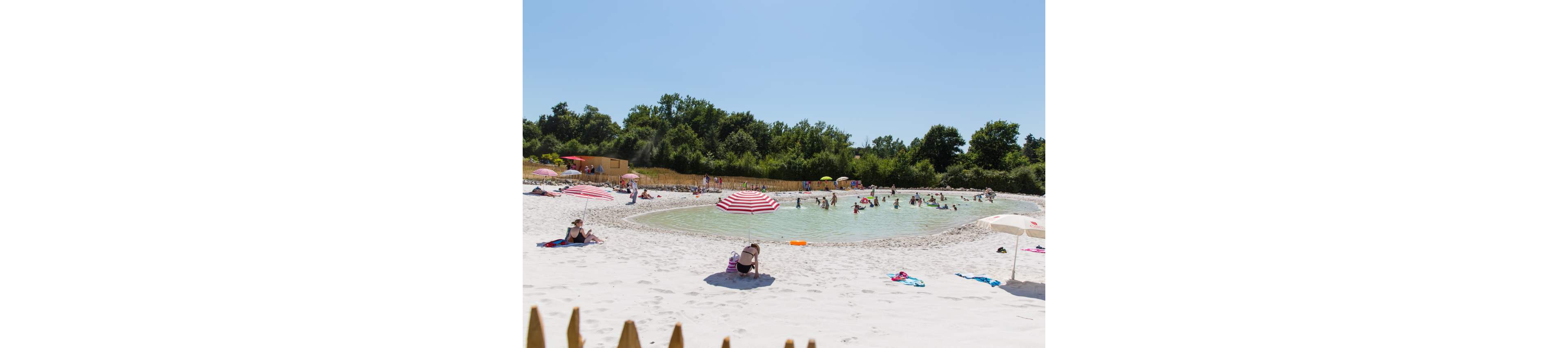 A man-made lagoon with pale turquoise water is surrounded by bright white sand, where many people relax and play. Families and children wade and swim in the shallow water while others sunbathe on towels and loungers. Colorful umbrellas, especially red-and-white striped ones, dot the shoreline and provide shade. Lush green trees and a clear blue sky frame the recreational beach setting, emphasizing a warm summer atmosphere.