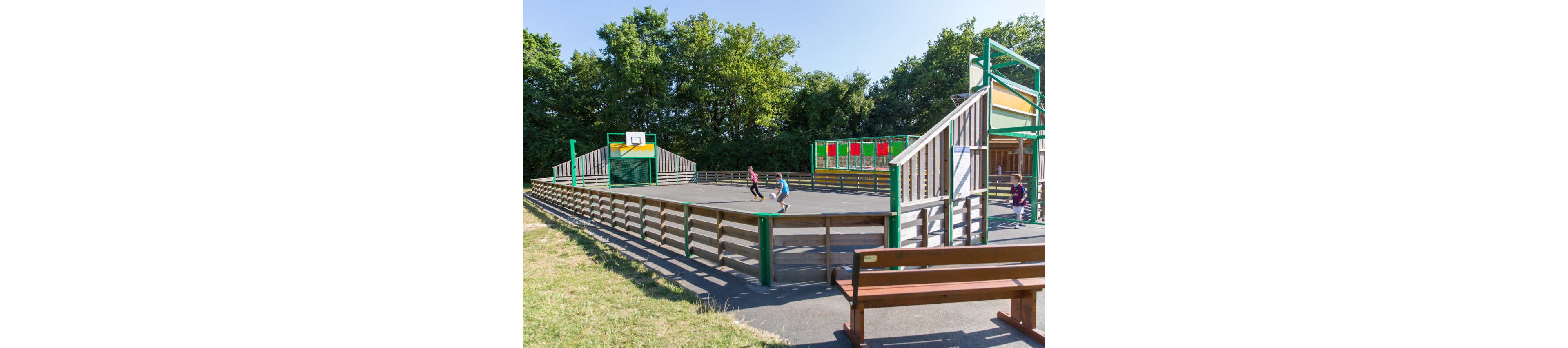 An outdoor multi-sport court features children playing basketball on a sunny day. The fenced court is surrounded by wooden barriers and colorful panels in green, yellow, and red. A basketball hoop stands at one end while benches line the paved area outside the court. Lush green trees form a natural backdrop, creating a bright and inviting recreational setting.