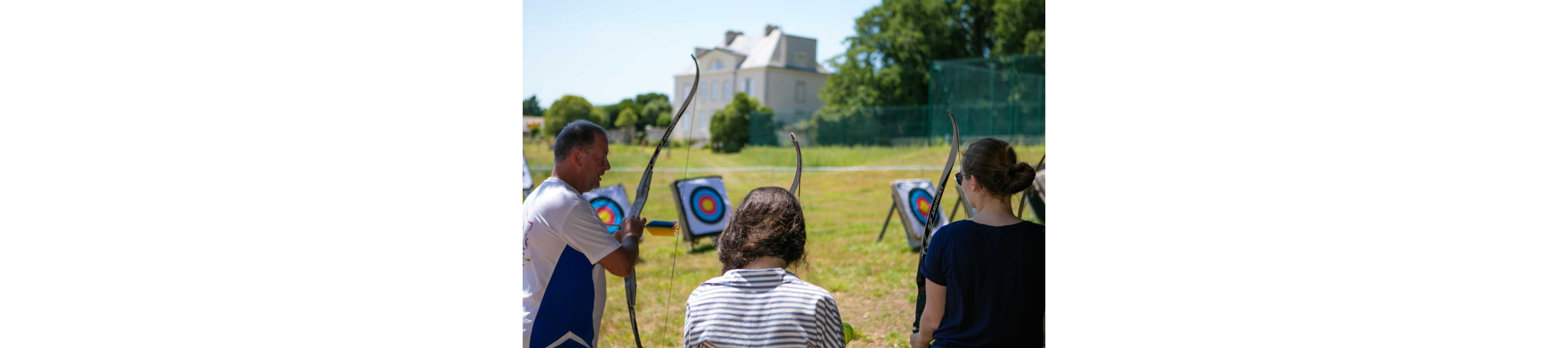 Three people stand on an outdoor archery range, each holding a bow and preparing to shoot. Colorful target boards are set up in the distance on a grassy field. A large house and trees are visible in the background under clear daylight. The scene suggests a recreational or training session in a relaxed, open-air setting.