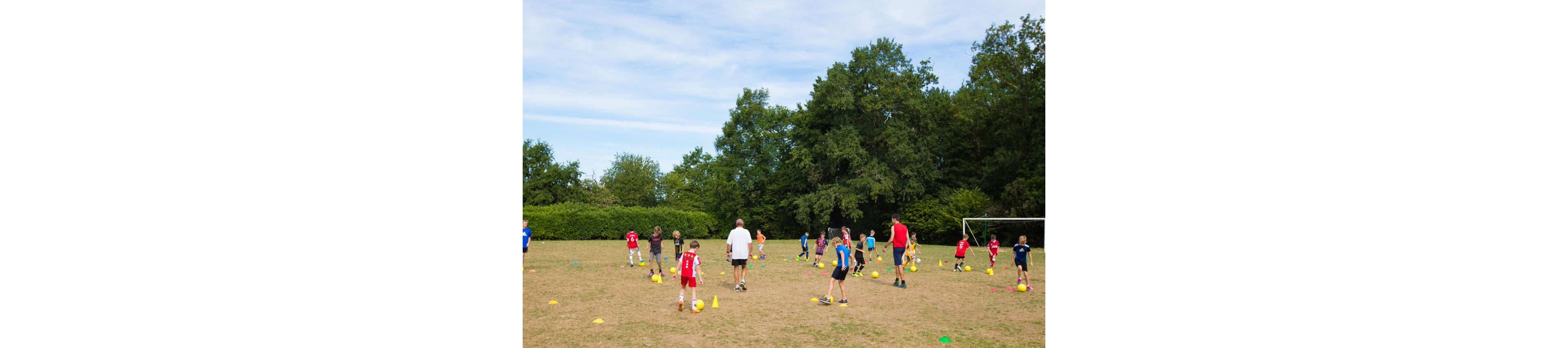 A group of children practice soccer drills on a grassy field. Several adults or coaches supervise as the kids dribble balls around colorful cones. The scene is set outdoors with trees and bushes surrounding the open space and a goalpost visible in the background. The atmosphere feels active and playful, suggesting a youth sports camp or training session.
