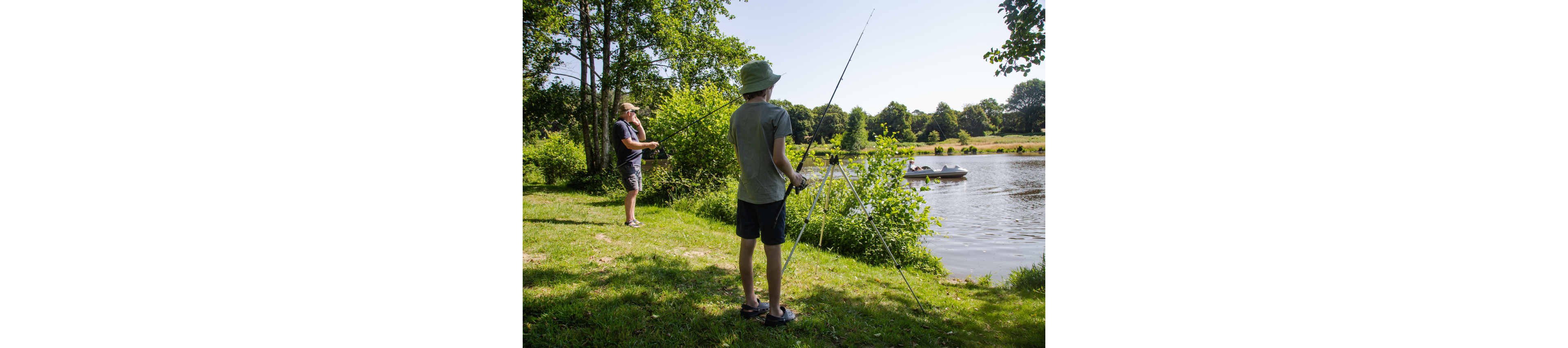 Two people are fishing on a grassy riverbank under bright summer sunlight. One person stands closer to the camera with a fishing rod and tripod, while another casts a line near the water’s edge. A small boat with people aboard moves along the calm river in the background. Lush green trees and open fields create a peaceful, natural setting.