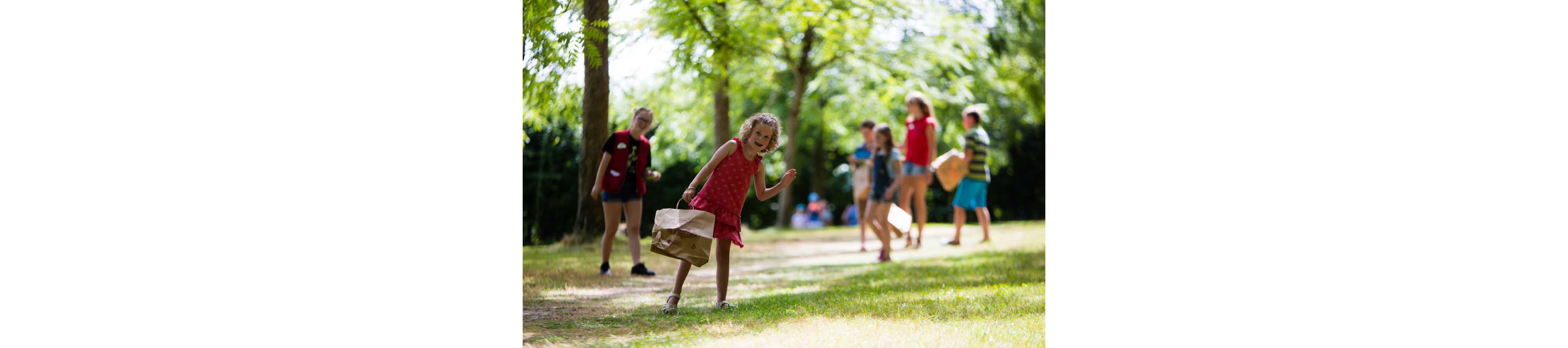 A group of children play an outdoor game in a sunny park. One child in the foreground bends forward while holding a brown paper bag, appearing focused on the activity. Other children stand and walk along the grassy path, also carrying paper bags. Tall green trees provide shade and create a bright, cheerful atmosphere. The scene suggests a summer camp or group activity in nature.