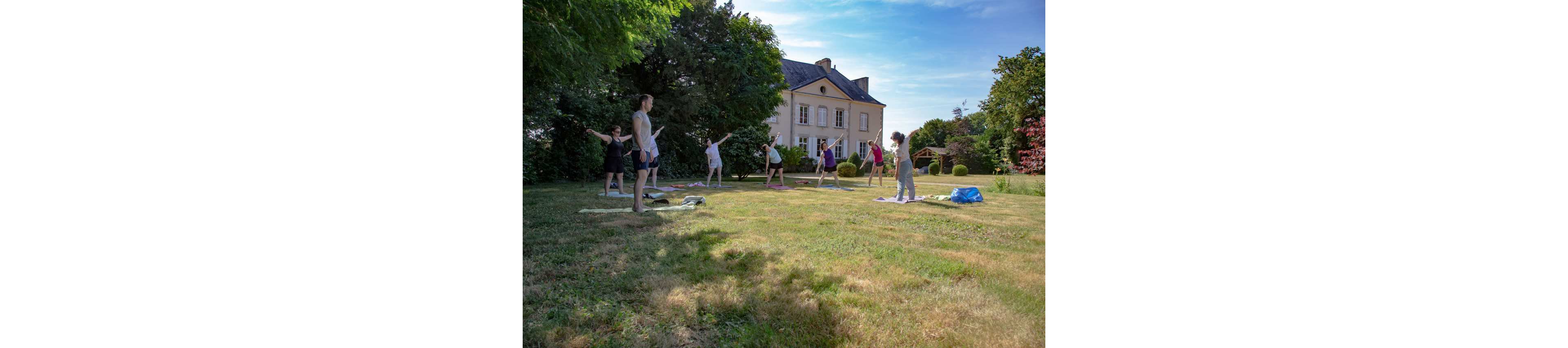 A small group of adults practices yoga on a grassy lawn in front of a large house. Participants stand on mats, stretching with arms extended in a coordinated pose. The scene is set in a sunny garden with trees, shrubs, and a well-kept yard. The bright daylight and greenery create a calm, relaxed atmosphere suitable for wellness and retreat activities.