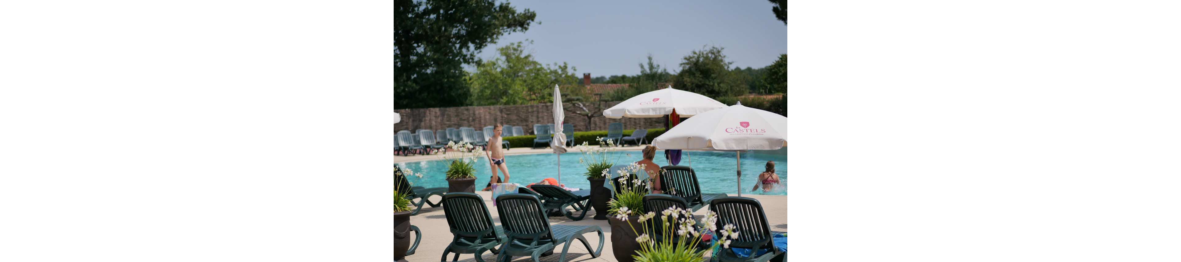 An outdoor swimming pool is surrounded by green lounge chairs and white parasols on a sunny day. Adults and children enjoy the water while others sit or recline near the poolside. Flowering plants in pots decorate the foreground, adding greenery and white blossoms to the scene. In the background, rows of empty loungers and trees suggest a quiet resort or holiday park setting.