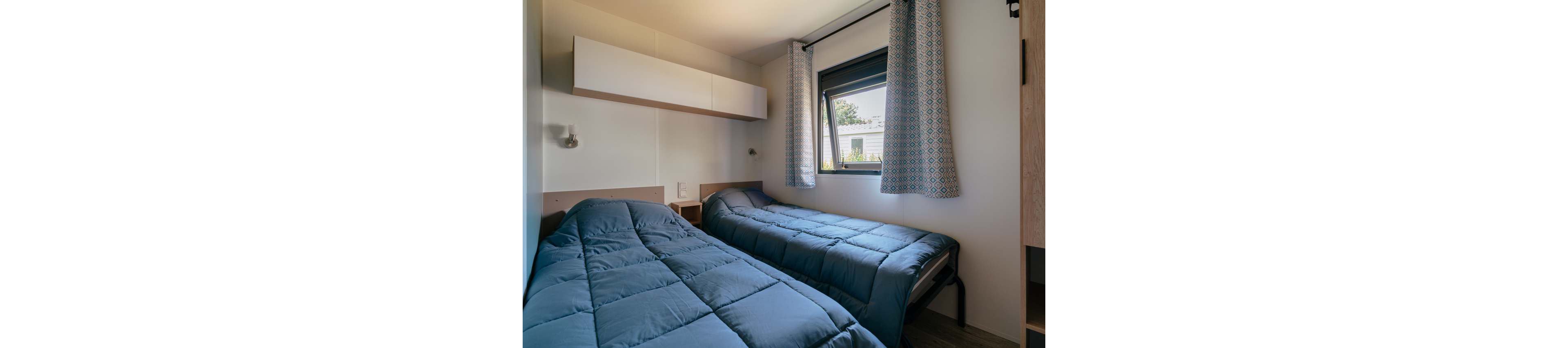A compact bedroom features two twin beds with blue comforters placed against opposite walls. Natural light enters through a single window with patterned curtains, revealing a view of neighboring buildings. Minimalist white cabinets and light wood accents create a clean, modern look. The room appears tidy and functional, suitable for guests or a vacation rental.