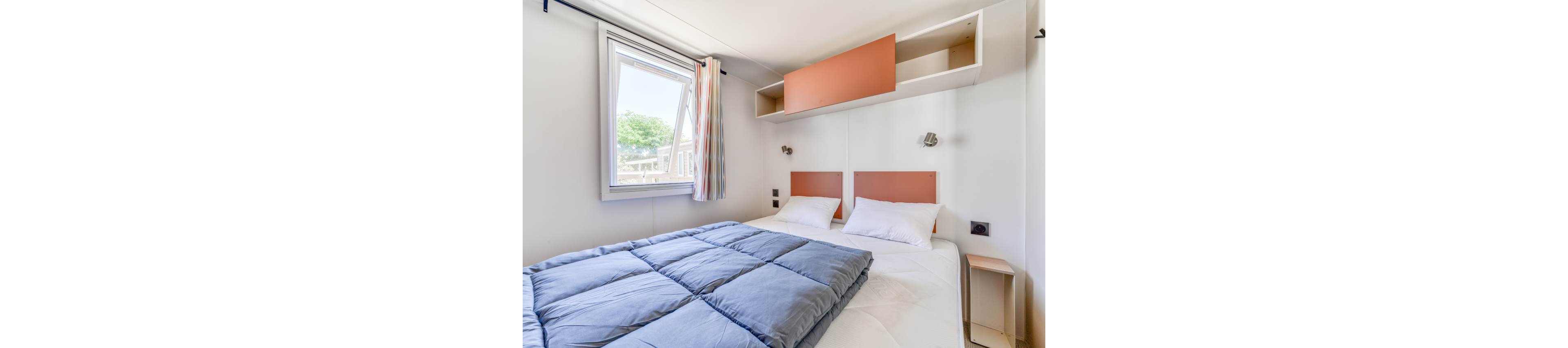 A small, modern bedroom features a neatly made double bed with a blue comforter and white pillows. The room has white walls with two rectangular headboards in a muted coral color. Built-in storage cabinets and shelves are mounted above the bed, adding functionality to the compact space. Natural light enters through an open window with striped curtains, giving the room an airy and inviting feel.