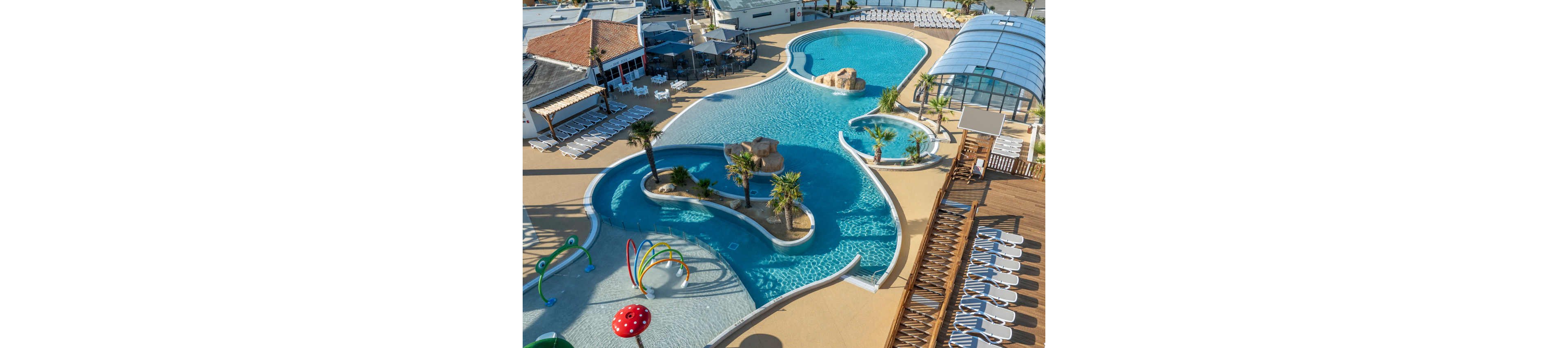 A large resort-style swimming pool complex is shown from above on a sunny day. The scene includes winding pools, palm trees, and rock features surrounded by lounge chairs and sun decks. A covered pool area and a shallow children's splash zone with colorful water sprayers are visible. The overall atmosphere suggests a family-friendly vacation destination with ample space for relaxation and play.