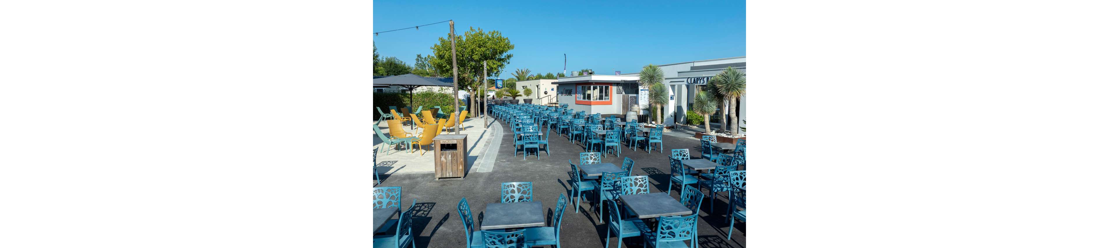 An expansive outdoor café terrace is filled with rows of blue metal tables and chairs, all currently empty. The setting appears to be a coastal or resort area, with palm trees and clear blue sky in the background. A small central building and surrounding structures house the café and adjacent businesses. Bright yellow chairs under a shaded area add contrast to the predominantly blue color palette. The scene conveys a calm, sunny day before guests arrive.
