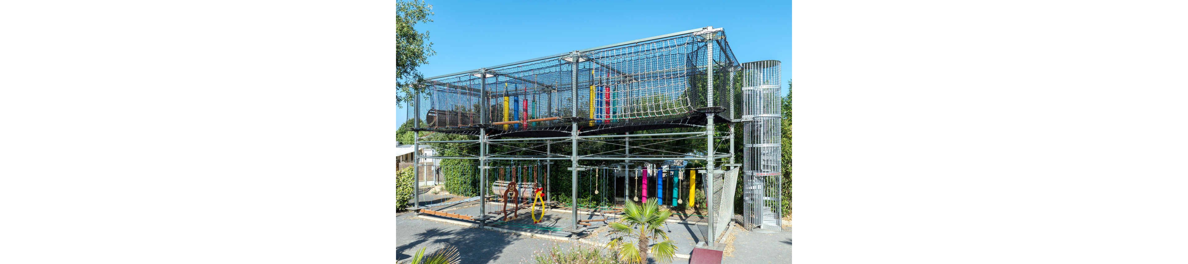 Large metal-framed adventure playground with elevated rope tunnels dominates the scene. The structure features multiple climbing elements, hanging obstacles, and colorful padded posts. It is set outdoors on a sunny day, surrounded by greenery and trees. The area appears to be part of a recreational park or campsite, with safety nets enclosing the upper level.