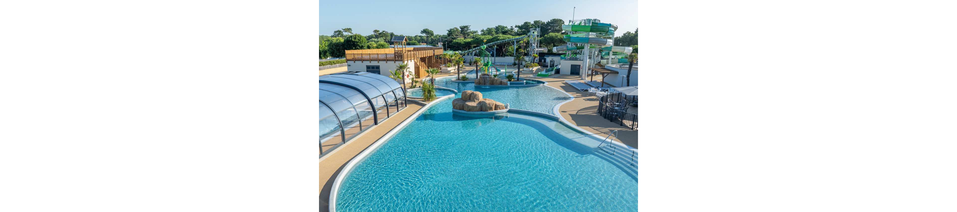A spacious outdoor water park features a large clear blue swimming pool with curved edges. In the background are multiple water slides, a splash play area, and a covered pool structure. Palm trees, rocks, and sun loungers add a resort-like atmosphere around the water. The scene is bright and sunny, suggesting a warm day ideal for family recreation.