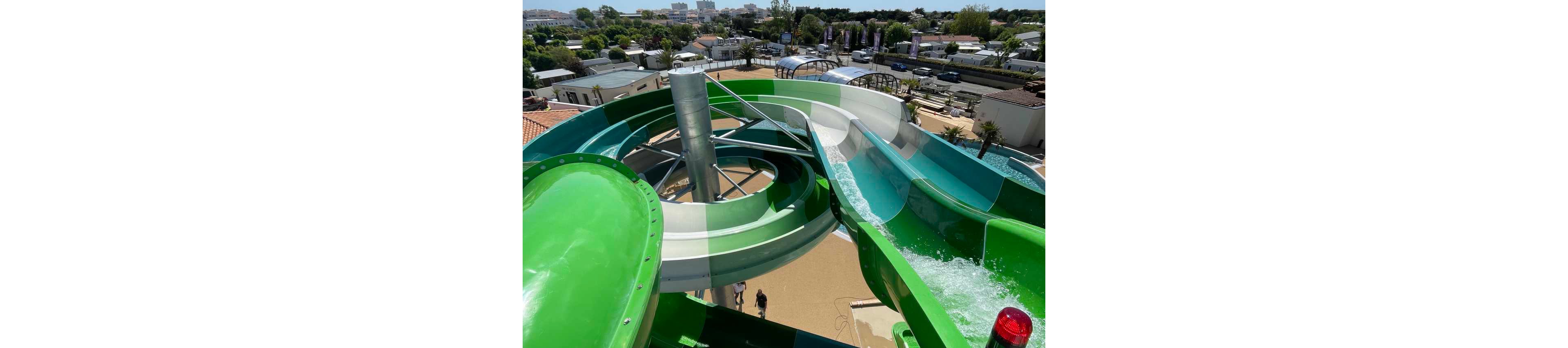 A large green and white waterslide twists around a central metal support structure at an outdoor waterpark. Sunlight reflects off the smooth slide surface as water flows down one of the chutes. Below, a few people walk on the tan ground near the base of the attraction. In the background, there are trees, buildings, and additional recreational facilities under a clear blue sky.