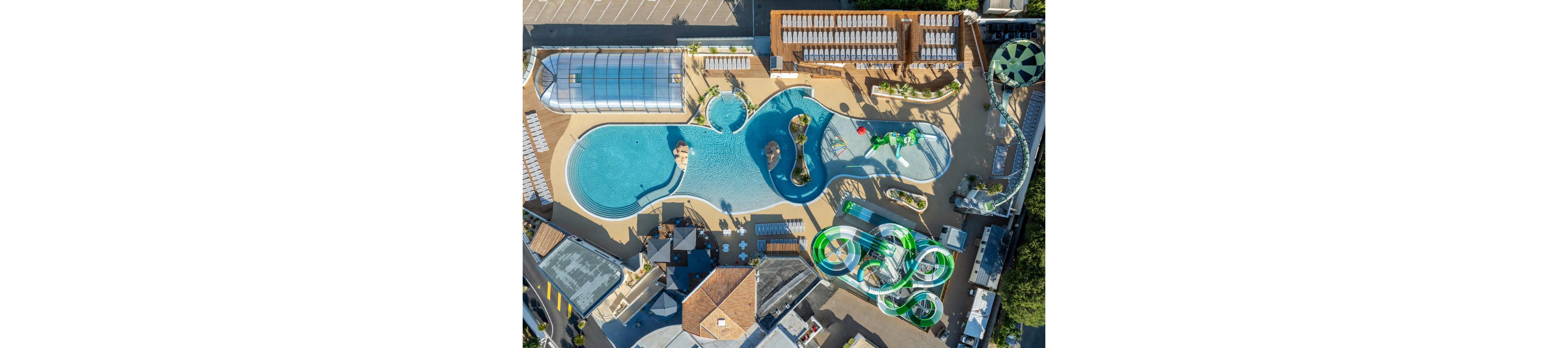 A large outdoor waterpark is seen from above, featuring expansive blue swimming pools and winding green waterslides. The complex includes sun loungers neatly arranged around the pools and several covered and open seating areas. Curved slides and splash structures create a playful contrast with the geometric layout of surrounding buildings and walkways. The overall scene suggests a sunny, leisure-focused environment designed for family recreation.