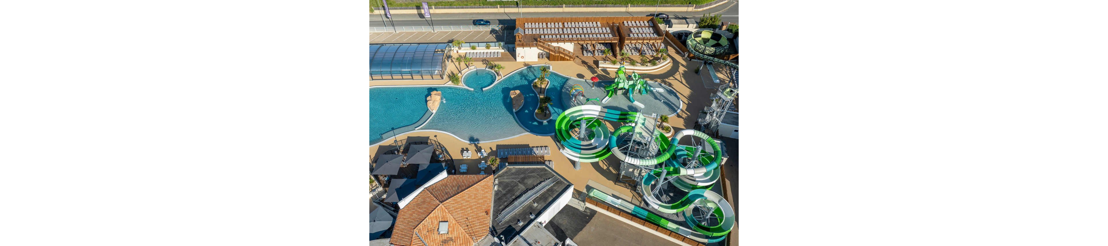 A large outdoor waterpark is captured from above, showing winding green and white waterslides and a spacious swimming pool. The setting includes sun loungers, shaded seating areas, and landscaped walkways around the pools. Nearby buildings, a covered pool structure, and a parking area with cars indicate a resort or recreational complex. The bright, sunny weather highlights the clean lines and vibrant colors of the water attractions.