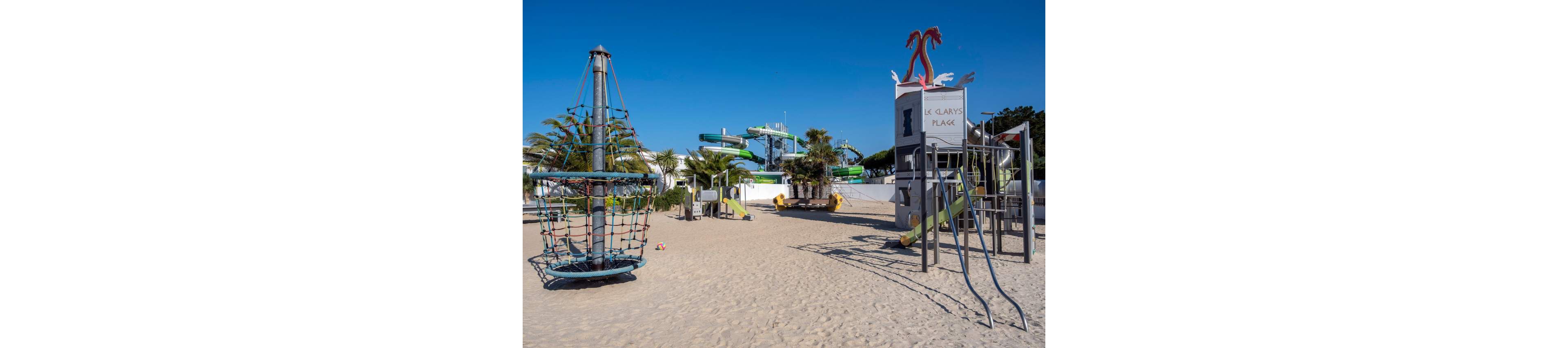 A sandy playground features climbing structures and rope equipment under a clear blue sky. In the background, a colorful water park with twisting slides rises above the trees. A tower structure with decorative seahorses displays the text "LE CLAYS PLAGE". The scene is bright and empty, suggesting a quiet moment at a family resort. Palm trees and metal play frames add to the coastal holiday atmosphere.