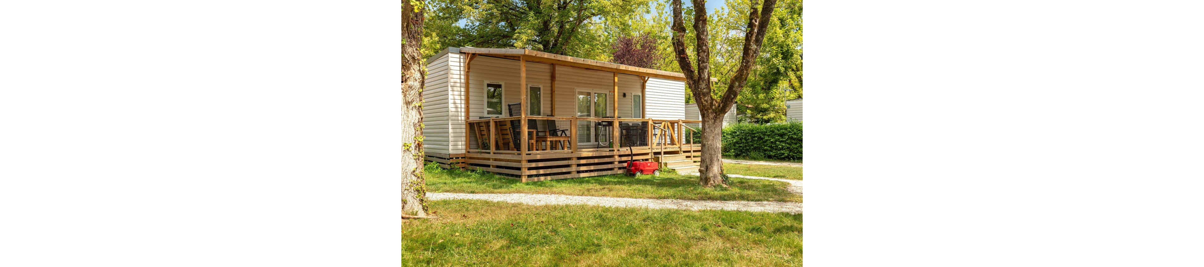 A modern mobile home with a spacious wooden deck sits in a green, tree-filled campsite. Outdoor chairs and a table are arranged on the covered terrace, suggesting a relaxing vacation setting. The lawn and gravel paths surround the unit, with hedges providing privacy between plots. Bright daylight and lush foliage create a warm, inviting holiday atmosphere.
