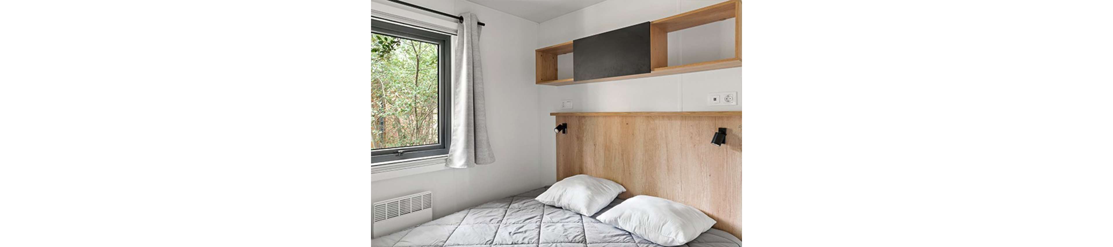 A small minimalist bedroom features a neatly made bed with a gray quilt and two white pillows. A light wood headboard with built-in shelving and a dark accent panel adds warmth to the white walls. A large window with gray curtains lets in natural light and offers a view of greenery outside. The overall design is clean, modern, and cozy, emphasizing simplicity and functionality.