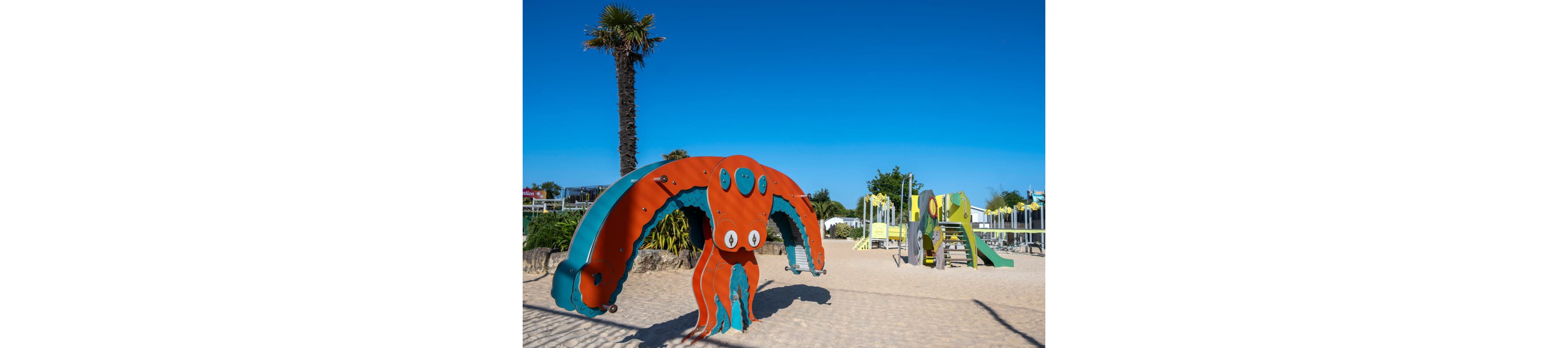 A brightly colored octopus-themed climbing structure stands on a sandy beach playground under a clear blue sky. In the background, additional play equipment and slides are visible, along with palm trees and low vegetation. The scene is sunlit, casting strong shadows on the sand and emphasizing the vivid orange, teal, and green play structures. The area appears calm and empty, suggesting an early or quiet time at a seaside recreation spot.