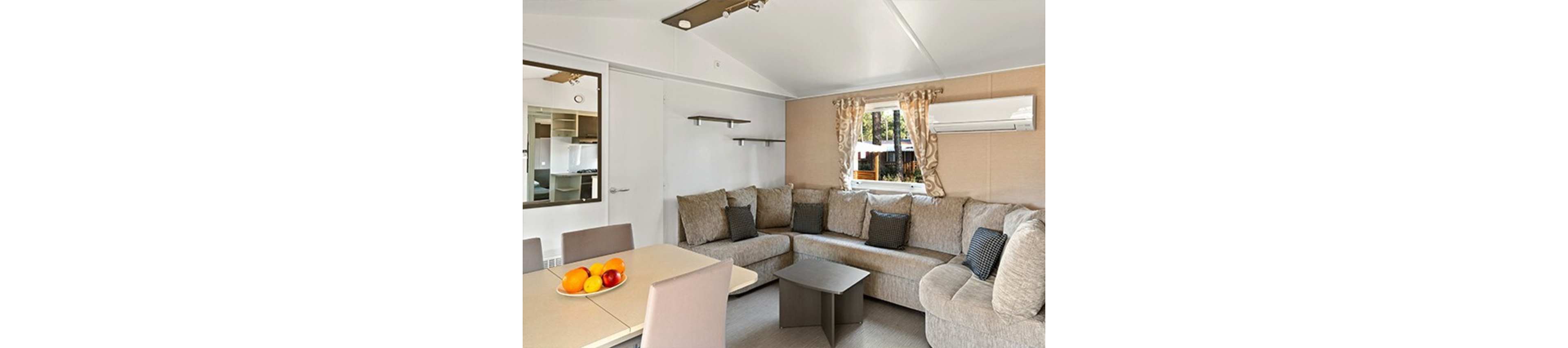 Bright living area featuring a large L-shaped beige sofa with multiple cushions. The space appears to be part of a compact vacation cabin or mobile home, with a dining table and chairs in the foreground. A small coffee table sits in front of the sofa, and an air conditioning unit is mounted on the wall above. Natural light enters through a window with patterned curtains, revealing trees and outdoor structures outside.