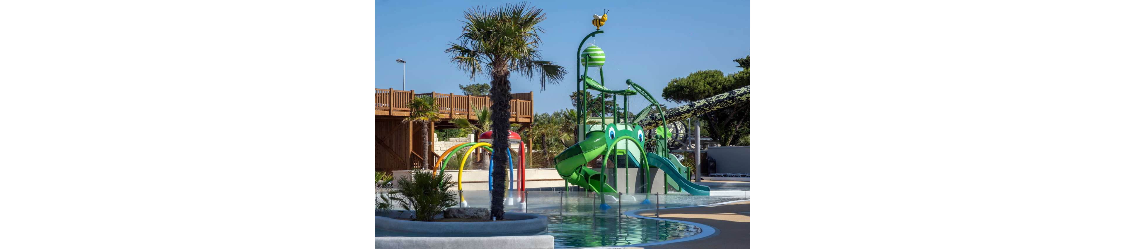 A vibrant outdoor water play area features a large green frog-themed slide structure with multiple chutes. The splash park is surrounded by shallow pools, curved walkways, and playful spray arches. Palm trees and lush greenery frame the scene, with a wooden deck and additional slides visible in the background. The clear blue sky and bright colors create a fun, family-friendly atmosphere.