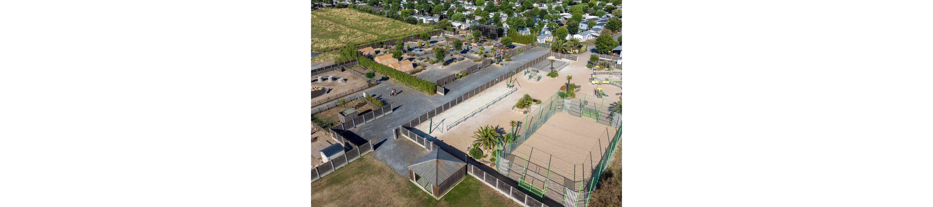 A large outdoor recreation complex is captured from an aerial perspective, showing multiple sports and play areas. The foreground features fenced sand courts and exercise equipment surrounded by palm trees and pathways. Additional play zones, walking paths, and landscaped sections extend into the distance. Beyond the complex, a residential or holiday park with numerous small homes and green spaces is visible.