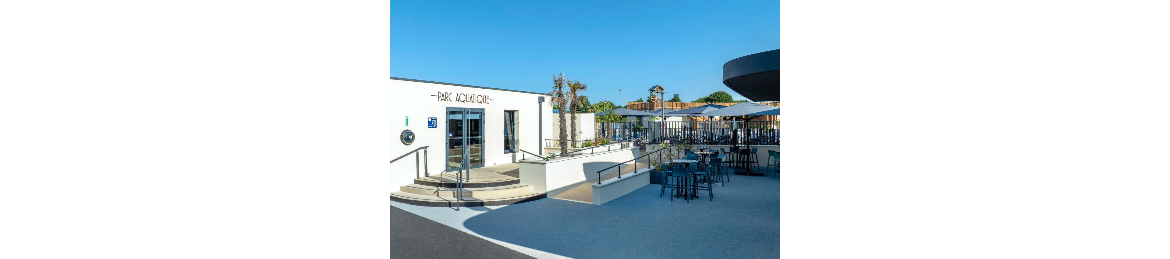 A modern white building marked 'Parc Aquatique' stands at the edge of a spacious outdoor terrace. The scene features tables and chairs arranged under umbrellas, suggesting a relaxed seating or café area. Palm trees, railings, and ramps add to the contemporary design of the aquatic park entrance. Clear blue sky and bright daylight create a clean, inviting atmosphere.