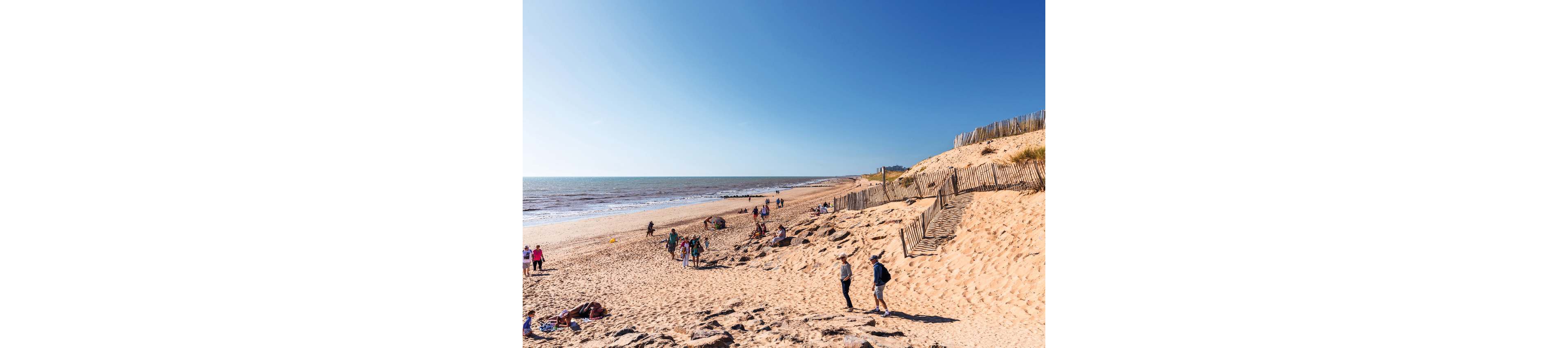 A wide sandy beach stretches along the coastline under a clear blue sky. Small groups of people walk, sit, and relax near the water and along the dunes. Wooden fences line the sand dunes on the right side, adding structure to the natural landscape. Gentle waves roll in from the sea, creating a peaceful seaside atmosphere.