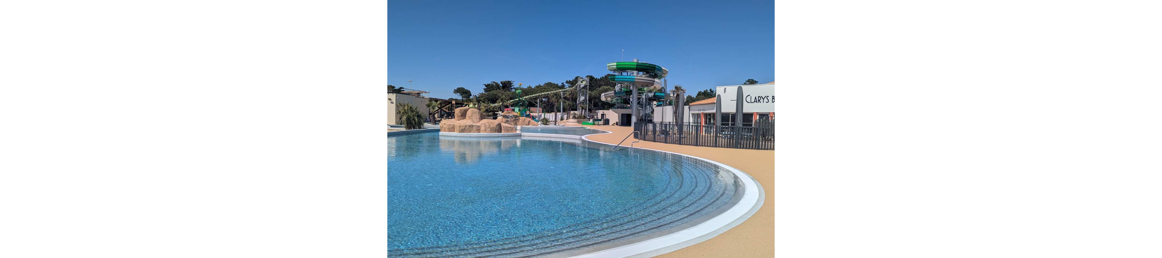 A large, clear blue swimming pool sits in the foreground of an outdoor water park. In the background, tall green water slides twist above a structure with stairs and platforms. Artificial rocks and landscaped areas add a natural touch around the pool. A nearby building with the visible text "CLARYS" suggests the name of the location. The scene is bright and sunny, with no people present, emphasizing a calm and inviting atmosphere.