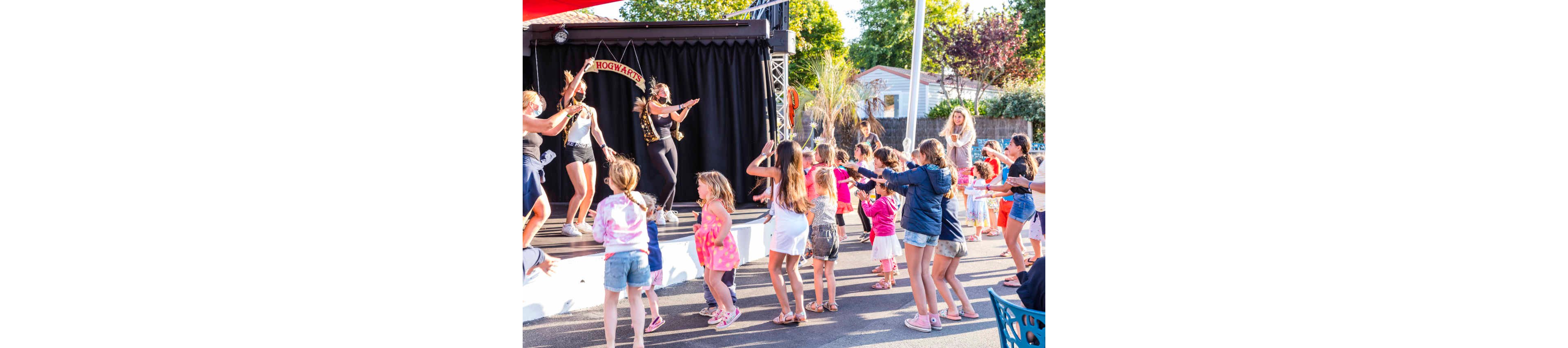 A group of children dance together in front of an outdoor stage during a lively performance. Several performers on the stage lead the moves, wearing costumes and headpieces under a small banner that reads "HOGWARTS." The scene takes place in a sunny open area with trees, buildings, and spectators in the background. Bright clothing and warm daylight create a cheerful, festive atmosphere.
