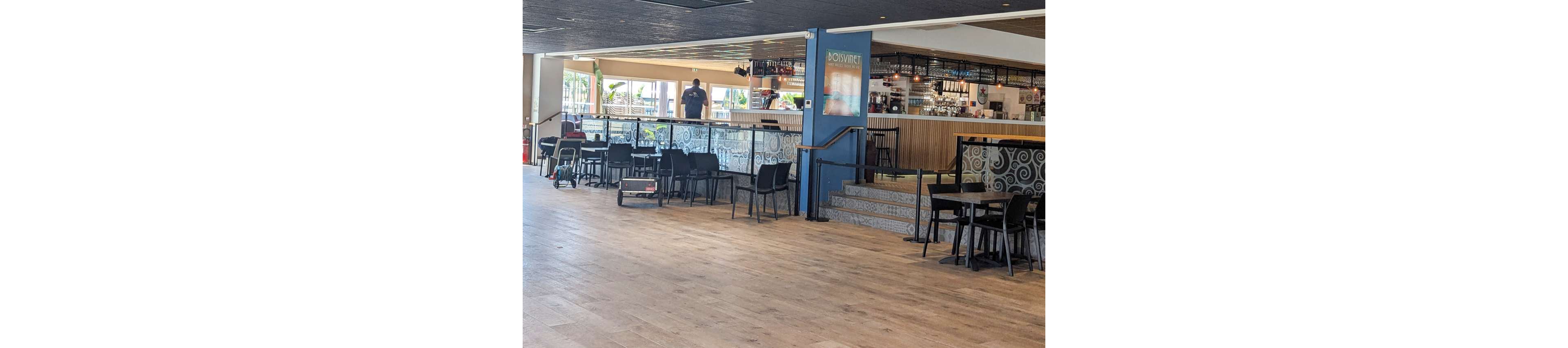 A large indoor bar area features rows of empty tables and chairs on a light wooden floor. The bar counter in the background is well lit, with bottles and glassware visible on the shelves. A single person stands behind the bar, adding to the quiet, relaxed atmosphere. Decorative tiles and a framed poster on a blue column add visual interest to the contemporary interior.