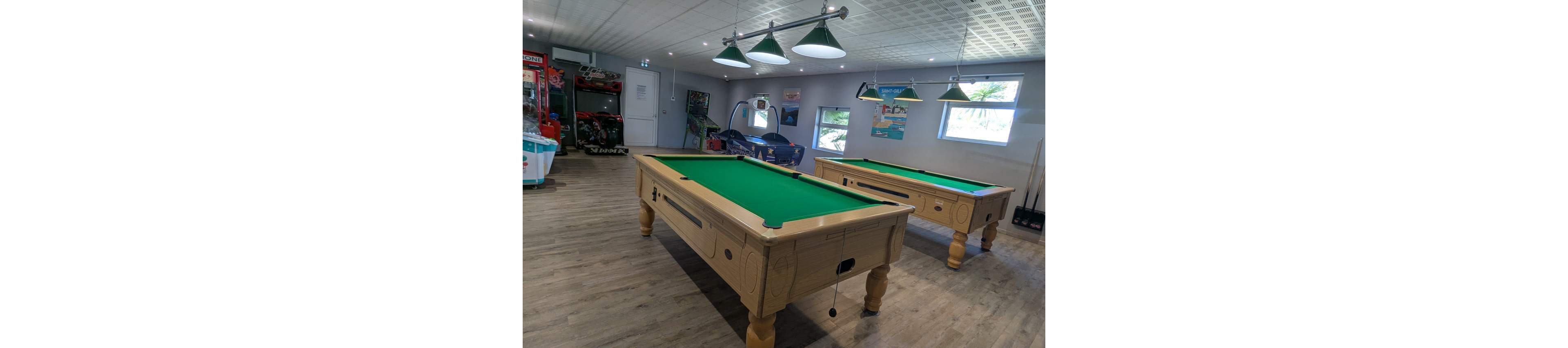 A bright indoor game room features two wooden pool tables with green felt tops as the main focus. The space has a light wood floor, white ceiling with recessed lighting, and large green hanging lamps above each table. In the background, various arcade machines and racing game simulators add to the entertainment atmosphere. Windows and wall posters provide additional color and visual interest around the room.