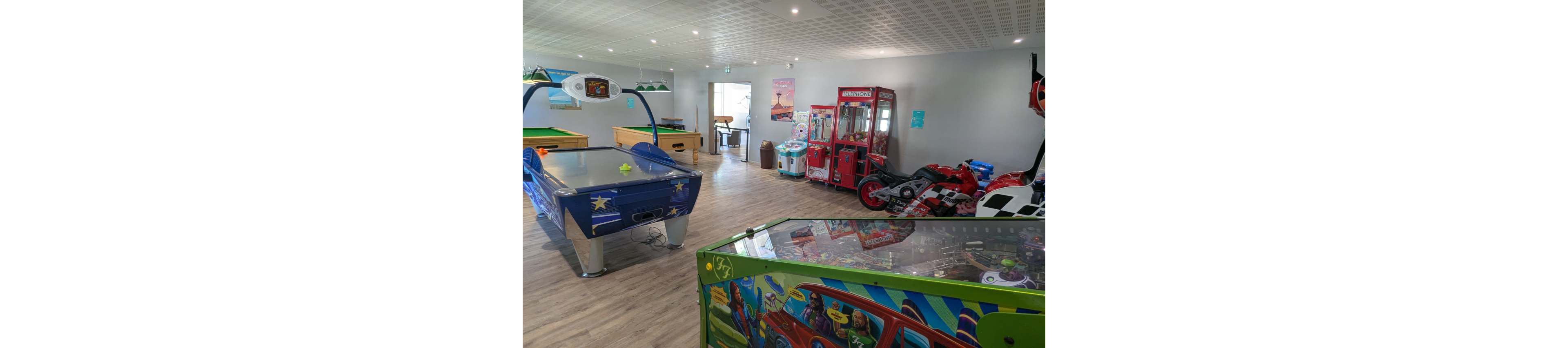An indoor arcade room features a variety of game machines, including an air hockey table, pinball machine, and racing motorcycle simulators. In the background, a claw crane machine and other arcade cabinets line the wall. The space has light-colored wooden flooring, white walls, and a modern ceiling with recessed lighting. Colorful graphics on the machines add a playful, family-friendly atmosphere.