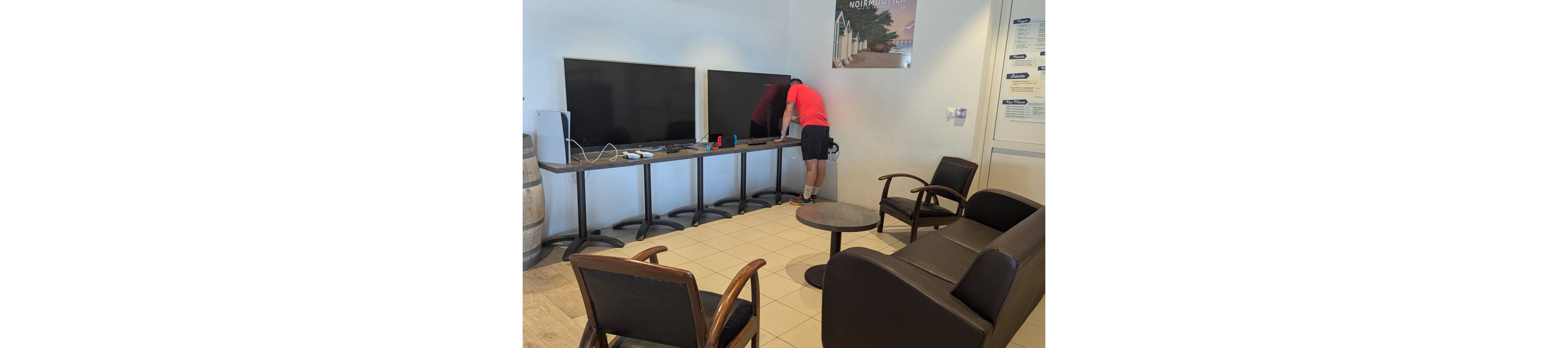 A man in a red shirt and black shorts adjusts cables behind large flat-screen monitors on a table. The scene takes place in a small waiting room with tiled floors and neutral-colored walls. Several chairs and a dark sofa surround a round coffee table in the foreground. Posters and informational notices are visible on the walls, adding to the functional, everyday atmosphere.