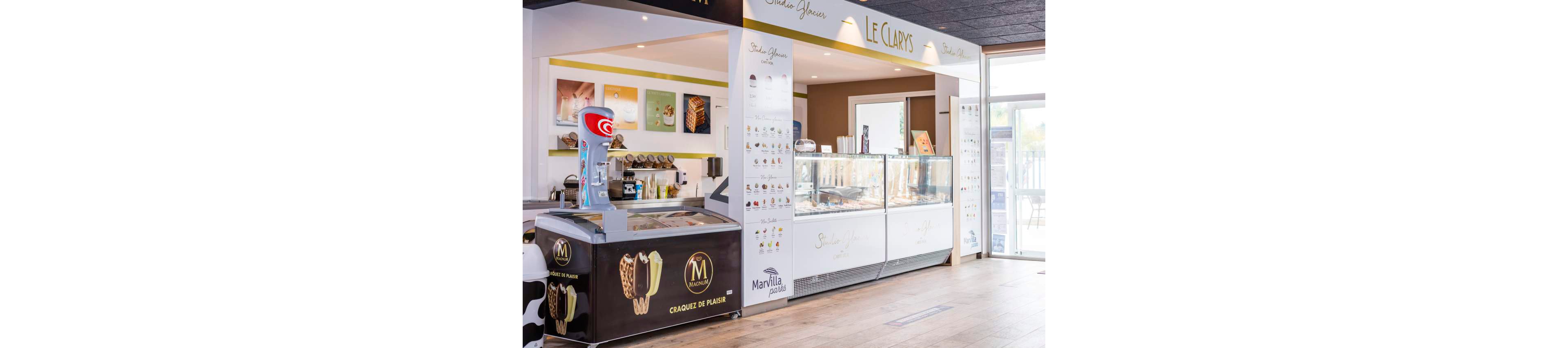 A bright ice cream parlor features large glass display freezers filled with frozen treats. A separate freezer counter in the foreground shows images of chocolate-coated ice cream bars. The interior has clean white walls, menu boards with dessert photos, and wooden flooring. Natural light enters from large windows on the right, giving the shop an inviting, contemporary feel.