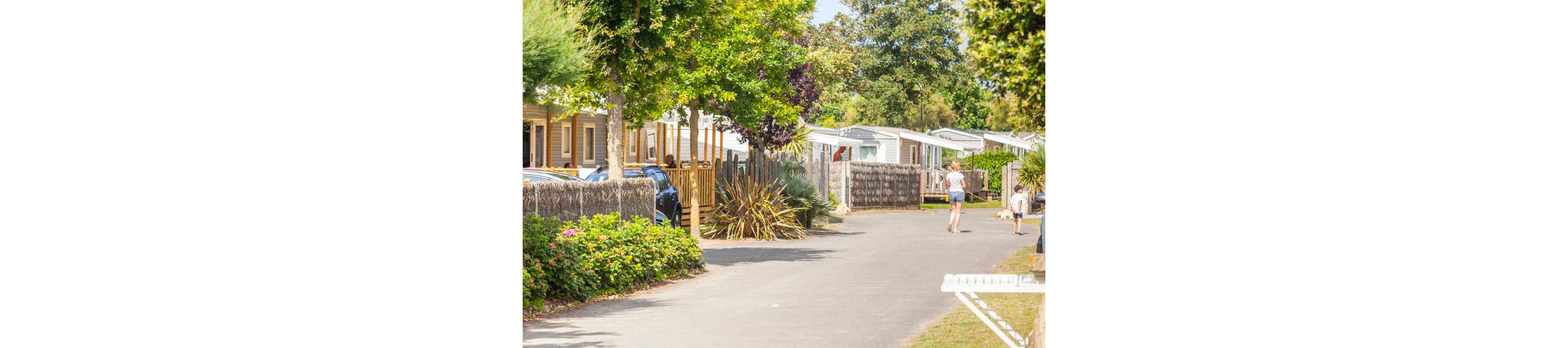 A quiet lane runs through a sunny mobile home park lined with trees and shrubs. Several light-colored caravans or mobile homes stand on either side, some with wooden decks and fences. A woman and two children walk along the paved path, suggesting a relaxed holiday atmosphere. Bright daylight and lush greenery create a warm, summery feel in this residential vacation setting.