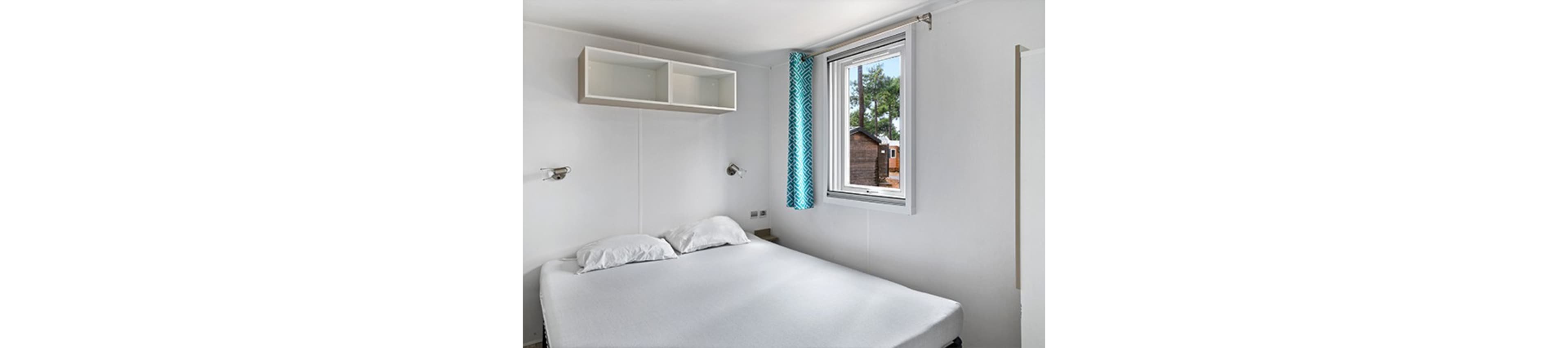 A small minimalist bedroom features a neatly made double bed with white bedding and two pillows. The walls and ceiling are white, enhancing the bright and clean appearance of the space. A window with a blue patterned curtain lets in natural light and offers a view of trees and a wooden shed outside. Wall-mounted reading lights and an open wall shelf add simple, functional storage and lighting.