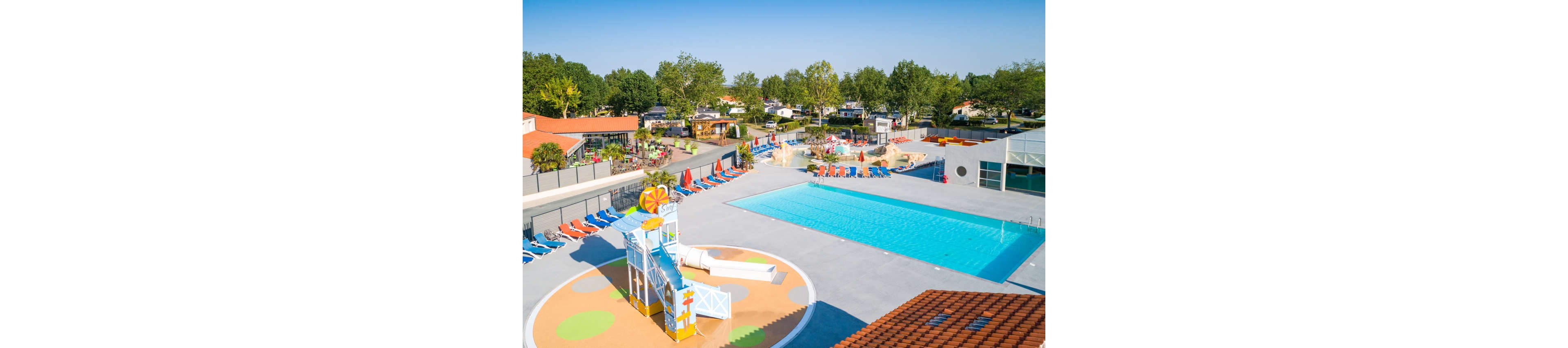 A large outdoor swimming pool is the central feature of this family resort. Colorful lounge chairs line the poolside, with a separate shallow play area featuring a small waterslide structure. The surrounding buildings and trees suggest a holiday park or campground setting. Bright sunlight and clear blue water create a cheerful, summery atmosphere ideal for relaxation and play.
