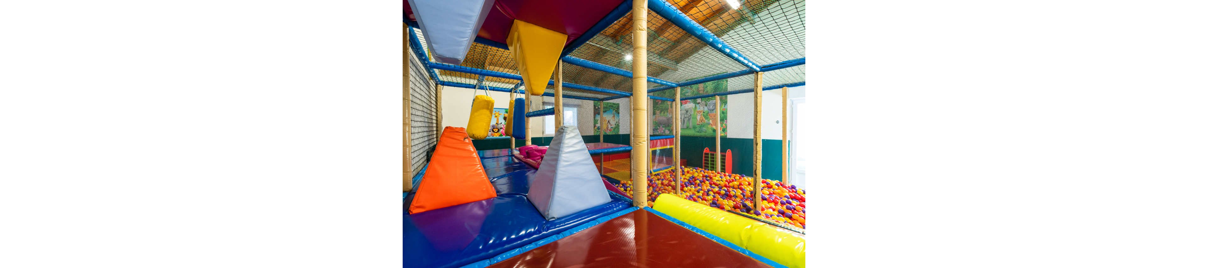 A bright indoor soft play area features padded climbing structures and ramps in vivid primary colors. The space includes netted walls and a safe, enclosed design suitable for young children. A large ball pit filled with multicolored plastic balls is visible in the background. The overall environment feels playful, energetic, and designed for active indoor fun.