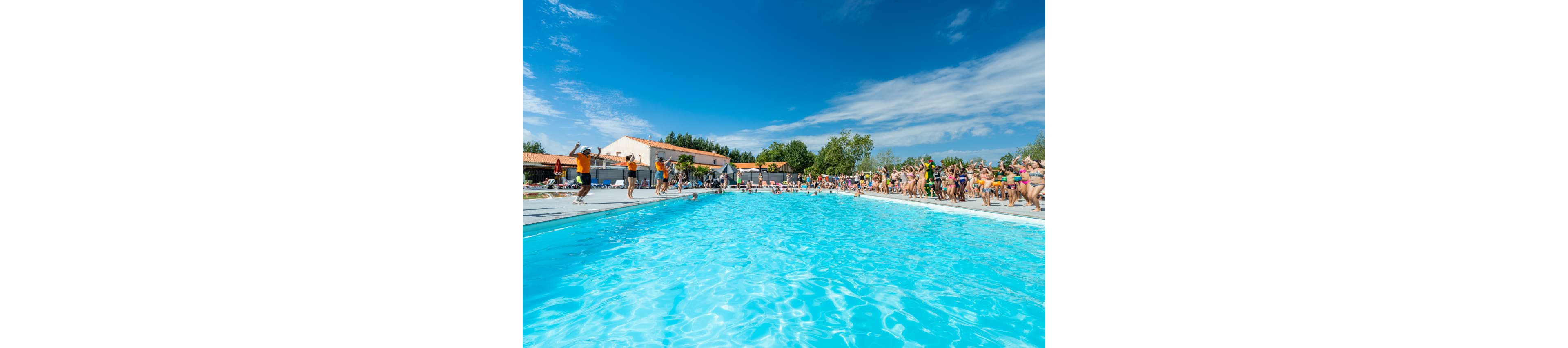 A large outdoor swimming pool is filled with bright blue water at a busy resort. Many people gather along the poolside, some standing and others preparing to jump into the water. Low buildings with orange roofs and surrounding trees frame the scene under a vivid blue sky with scattered clouds. The atmosphere feels festive and energetic, suggesting a summer vacation or holiday event.
