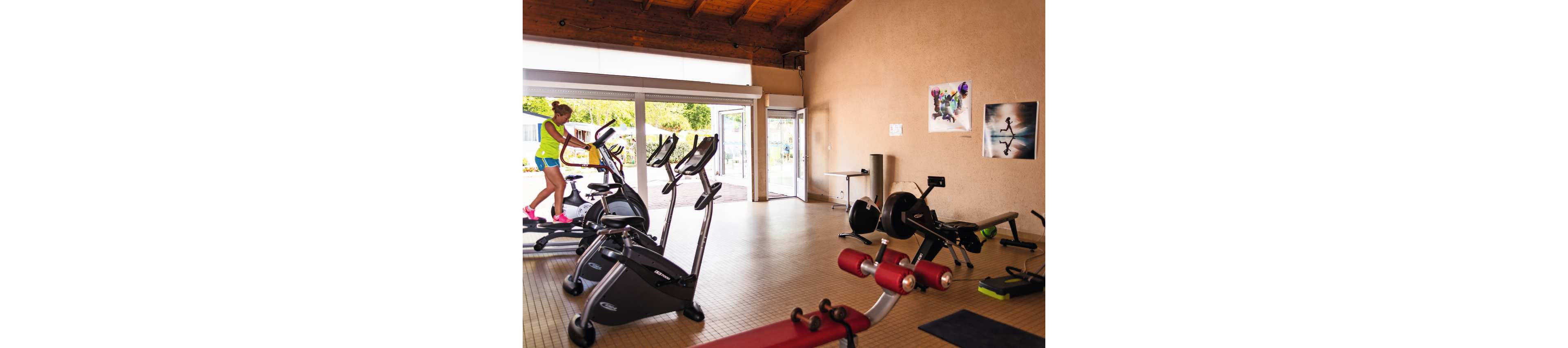 A woman works out on a cardio machine in a small, well-lit gym. Several exercise bikes and fitness machines are lined up facing large open glass doors. Natural light streams in from outside, creating a bright and airy atmosphere. Posters of athletic figures decorate the beige walls, and red and black equipment is arranged neatly on the tiled floor.