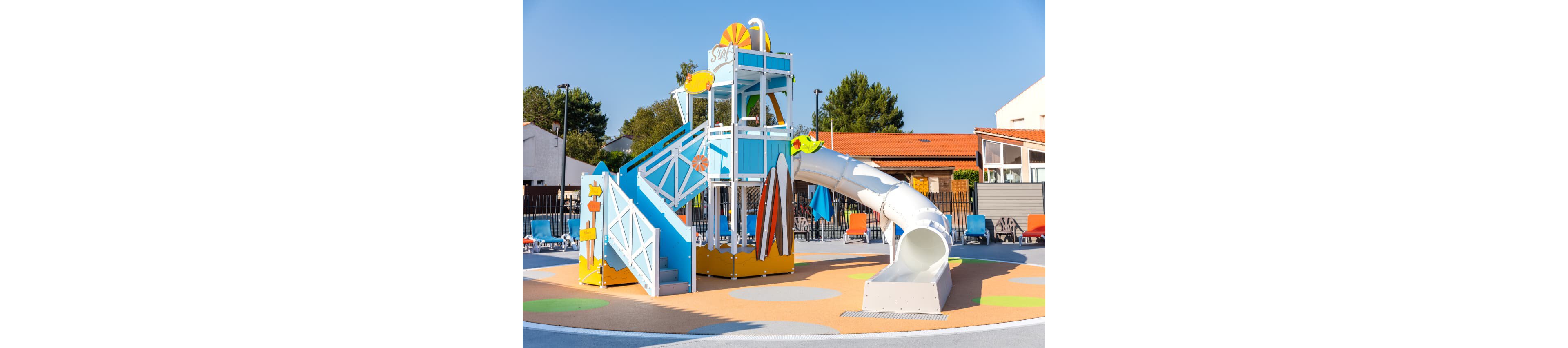 A bright, modern water playground structure stands in an outdoor recreational area. The play tower features multiple levels, surfboard decorations, and a large enclosed tube slide that curves down to the splash pad. The ground is covered with colorful, soft safety surfacing in circular patterns. In the background, there are lounge chairs, umbrellas, and buildings with red-tiled roofs under a clear blue sky.