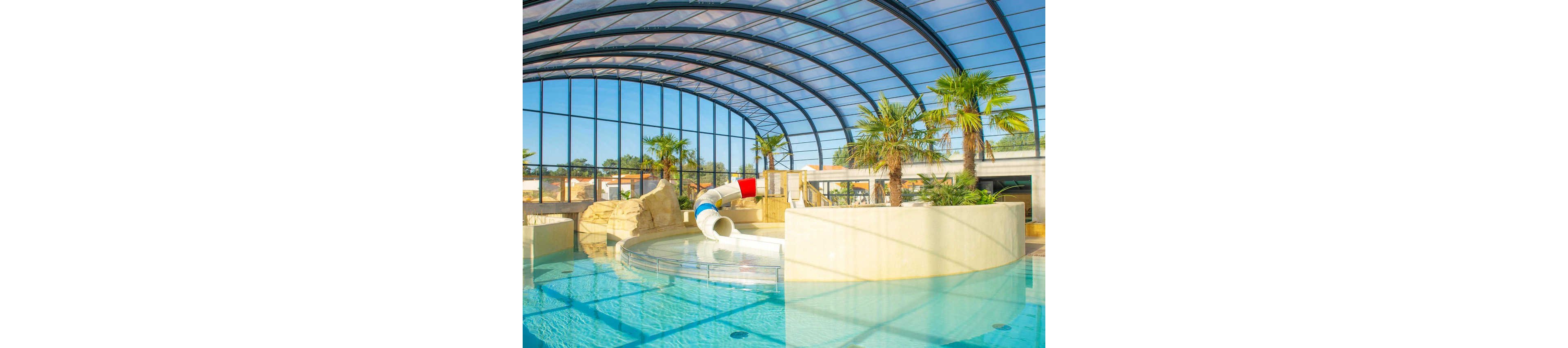 A bright indoor swimming pool features a curved waterslide ending in a shallow basin. The pool is housed under a large glass roof that lets in abundant natural light. Palm trees and rock-like structures create a tropical, resort-style atmosphere. Clear blue water and smooth beige walls give the scene a clean, modern look.