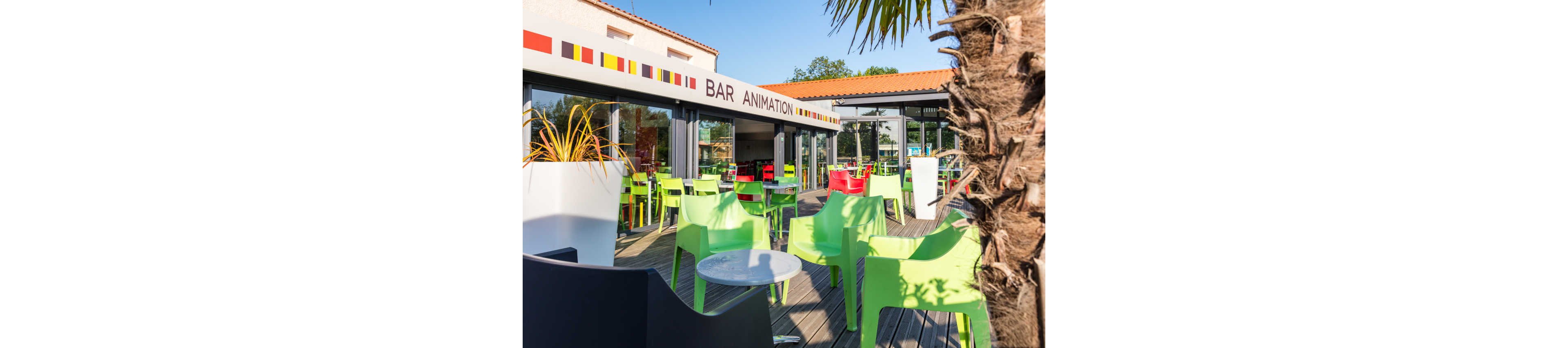 A modern outdoor bar terrace features bright green and black plastic chairs around small tables. The area is set on a wooden deck in front of a building with large glass doors and a red-tiled roof. A sign reading "BAR ANIMATION" is visible above the entrance, along with a decorative band of colored squares. Palm leaves and potted plants add a relaxed, holiday atmosphere. The scene is sunlit and appears ready for guests but currently empty.