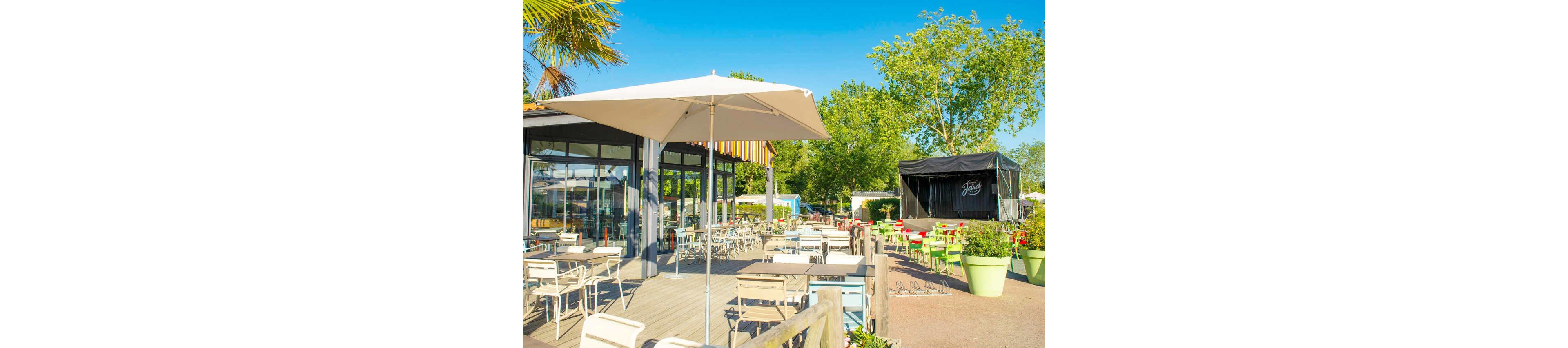 An outdoor café terrace is set with numerous tables and chairs under large white umbrellas. The setting appears to be a modern restaurant or bar with glass walls and colorful awnings. Bright green plants in oversized pots line the patio, adding a lively touch to the scene. A small stage structure is visible in the background, suggesting live performances or events. The overall atmosphere is sunny, relaxed, and inviting for outdoor dining.