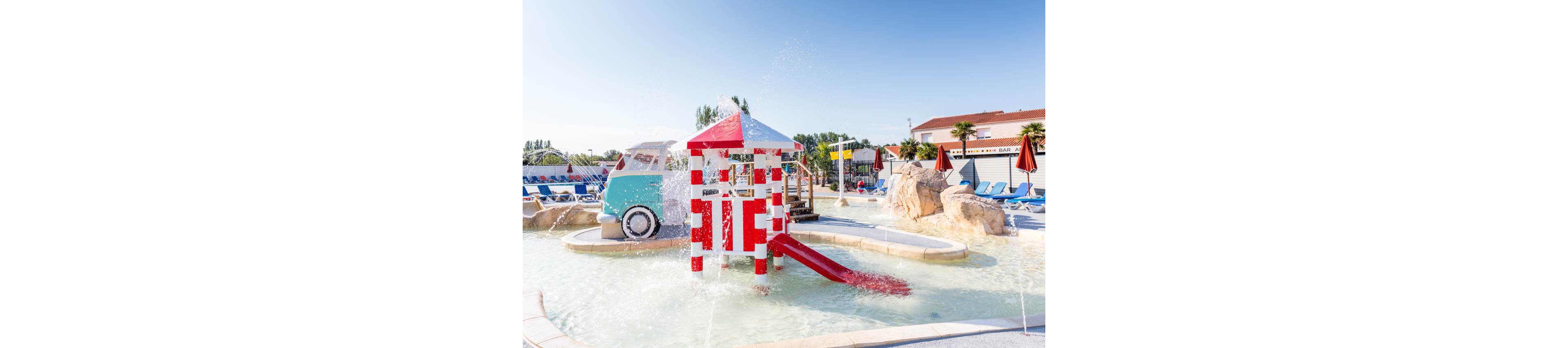A bright children’s splash park features a red and white play structure with a small water slide in a shallow pool. Water sprays from multiple fountains, creating a playful and refreshing atmosphere. In the background, a retro-style van structure and artificial rocks add visual interest. Rows of blue lounge chairs and umbrellas suggest a family-friendly resort or water park setting under a clear blue sky.