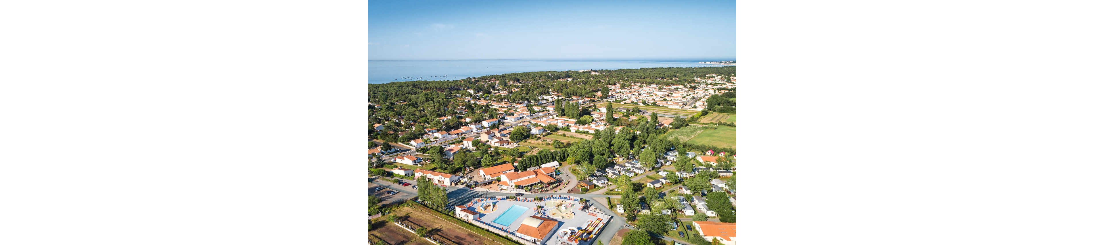 An aerial view shows a coastal town with a large campsite and swimming pool complex in the foreground. The scene includes multiple pools, recreational areas, and rows of buildings with red-tiled roofs surrounded by trees. Residential houses and caravans extend across the landscape, leading toward a distant shoreline and calm sea. The image captures a sunny day with clear skies and lush greenery throughout the area.