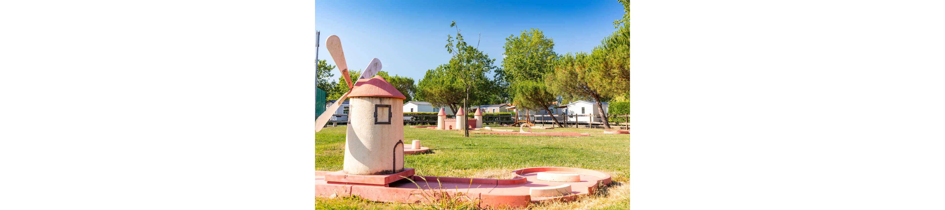 A small outdoor mini golf course features several windmill-shaped obstacles with red roofs and white bodies. The course is set on a grassy field under a clear blue sky, surrounded by trees. In the background, white mobile homes or cabins line the edge of the property, suggesting a campground or holiday park setting. The scene is bright and sunny, creating a cheerful and relaxed atmosphere.