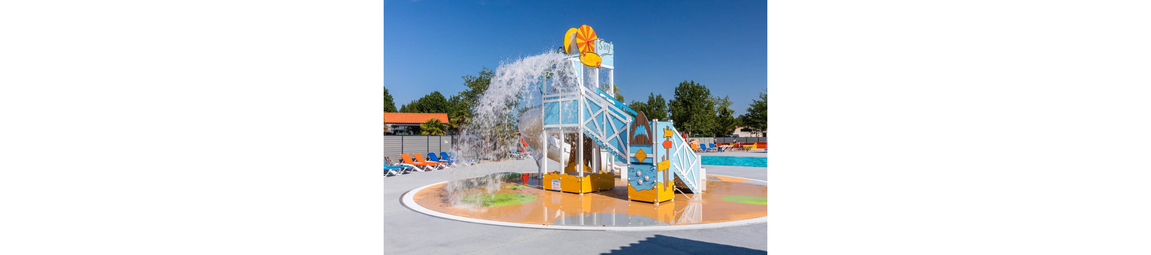 A bright water playground structure pours a large splash of water onto a shallow splash pad. The play equipment features slides, stairs, and decorative surfboard motifs in blue, white, and yellow. In the background, a large outdoor swimming pool and rows of colorful lounge chairs sit under a clear blue sky. Trees and low buildings frame the sunny recreational setting, creating a cheerful family-friendly atmosphere.