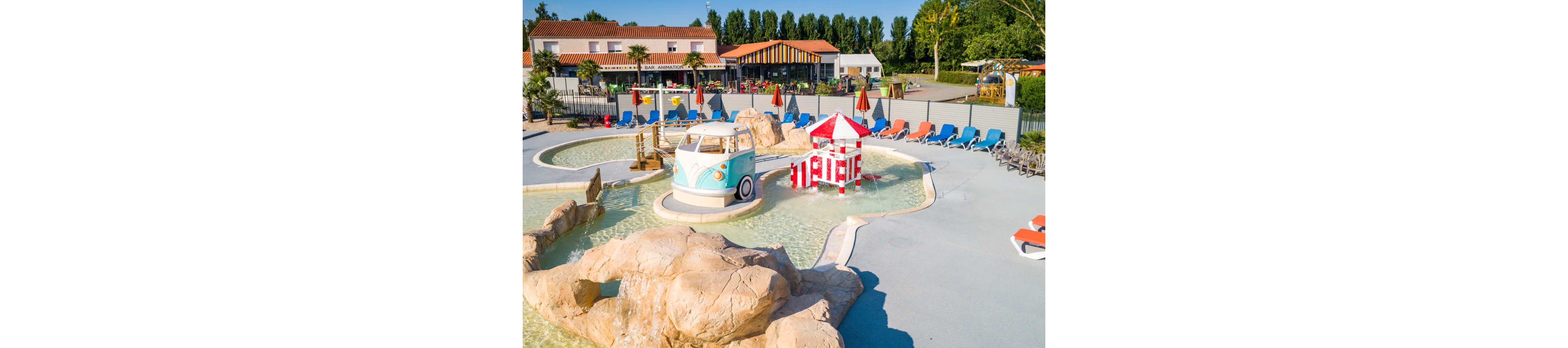 An outdoor children’s splash park features shallow pools with playful structures, including a retro van-shaped play unit and a red-and-white striped tower. The area is surrounded by sun loungers and umbrellas, suggesting a family-friendly water recreation space. In the background, low buildings and trees frame the leisure complex under clear skies. The bright colors and clean design create a cheerful, inviting atmosphere for kids and parents.