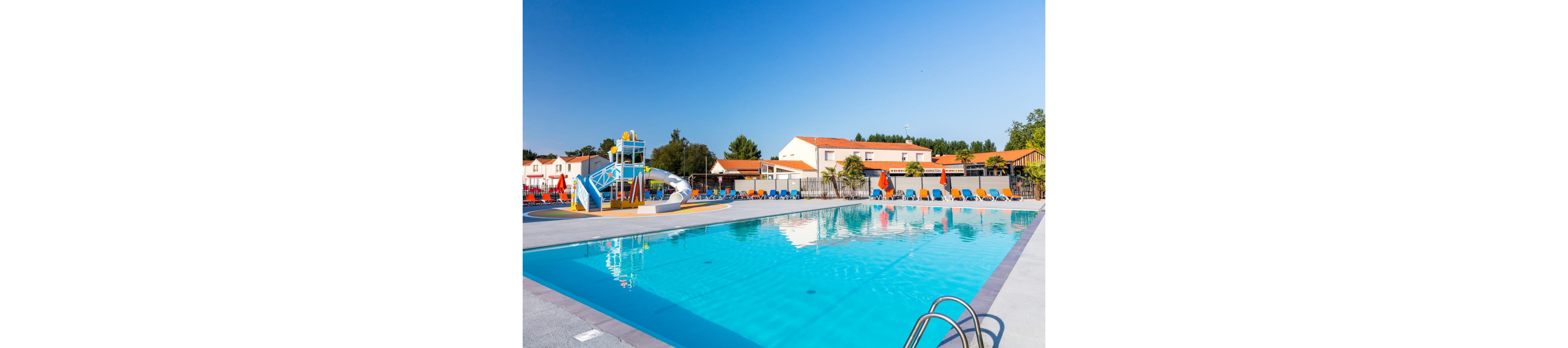 A large outdoor swimming pool is featured in a bright, sunny resort setting. Colorful lounge chairs line the poolside, facing the clear blue water. A small water play structure with slides and sprayers stands near the pool, suggesting a family-friendly environment. Low-rise buildings with red-tiled roofs and a clear blue sky complete the relaxed vacation atmosphere.