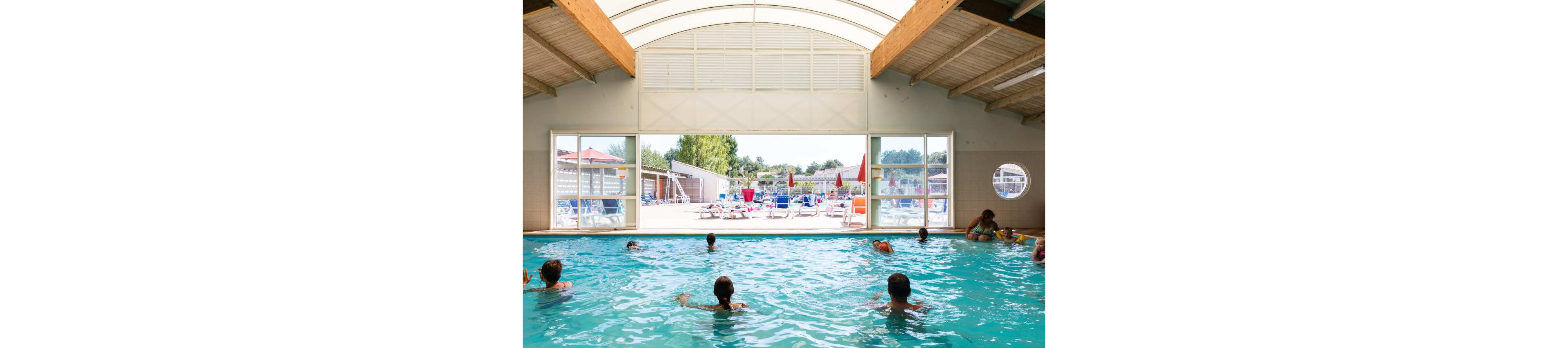 Several people swim and relax in an indoor swimming pool under a high wooden roof. Large open windows reveal an outdoor patio area with sun loungers and umbrellas in bright daylight. The scene suggests a leisure or vacation setting, possibly at a resort or campground. Cool blue water contrasts with the warm tones of the wooden ceiling and the sunny exterior.